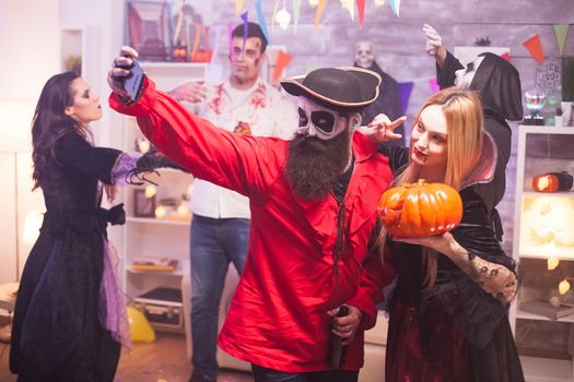 Beautiful vampire woman and her pumpkin taking a selfie with bearded pirate celebrating halloween.