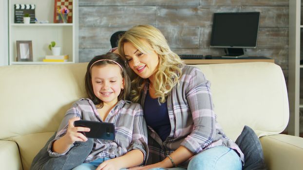 Young mother and her daughter relaxing on the couch watching a funny video on smartphone.