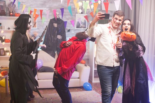 Male zombie taking a selfie with woman witch holding a pumpkin at halloween celebration.