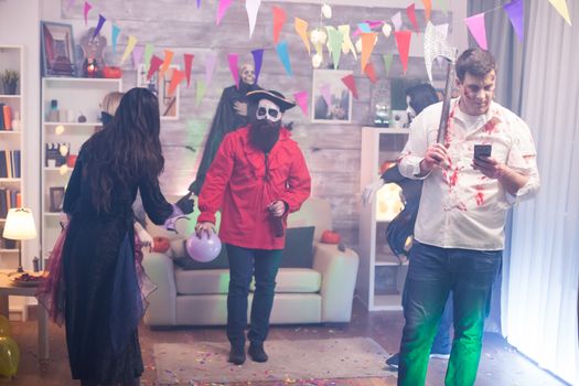 Young man dressed up like a zombie with an axe scrolling on his phone at halloween party.