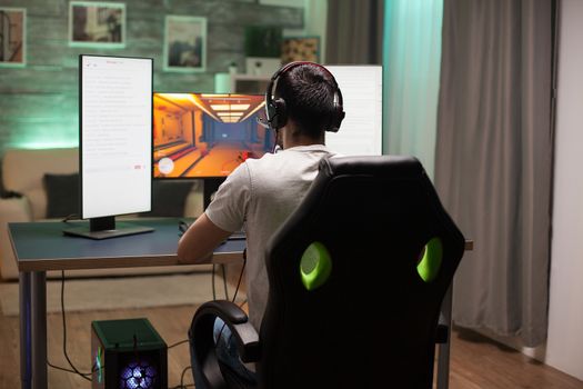 Back view of man sitting on gaming chair playing shooter game in the night. Neon light in the room.