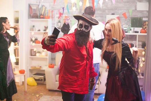 Blond vampire woman taking a selfie with a medieval pirate at halloween celebration.
