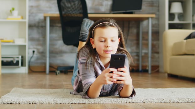 Cheerful little girl lying on the floor carpet using smartphone.