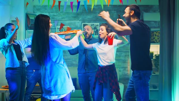 Attractive young woman arriving with tasty pizza at the party for her friends. Neon lights, alcohol and disco ball