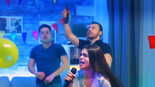 Beautiful young woman singing a song on microphone while partying with her cheerful friends. Karaoke at wild college party in room with neon lights, disco ball and alcohol