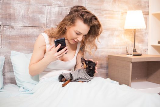 Relaxed beautiful woman in pajamas talking with her little dog sitting on bed at night.