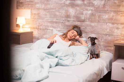 Beautiful caucasian woman falling asleep holding tv remote control. Little dog.