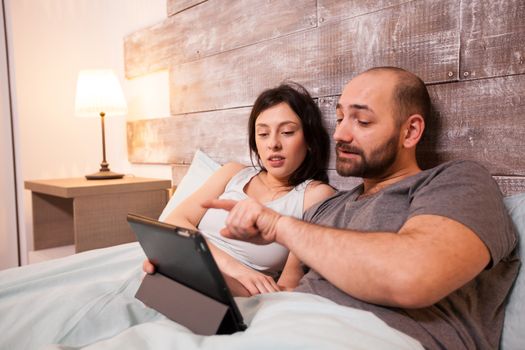 Beautiful couple wearing pajamas lying in bed surfing on internet using tablet computer.