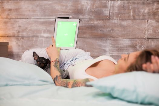 Beautiful woman with pajamas in bed relaxing using tablet computer with green screen. Cute little dog.