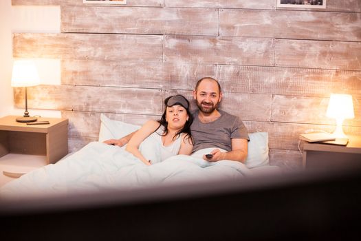 Young couple in pajamas watching tv lying under the blanket on comfortable bed.