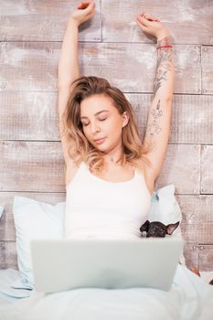 Sleepy beautiful woman in pajamas at night stretching while working on laptop.