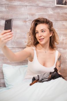 Sexy woman wearing pajamas taking a selfie with her dog in bed at night.