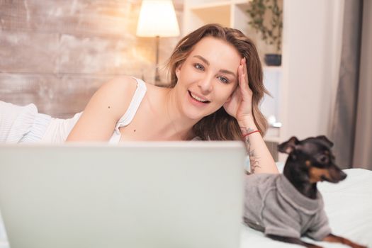 Little dog ignoring her beautiful owner wearing pajamas and using laptop.