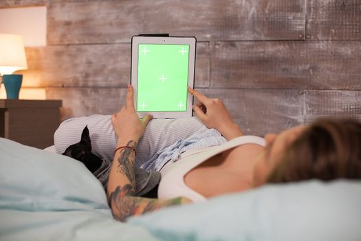 Young woman lying in bed wearing pajamas pointing at tablet computer with green screen.