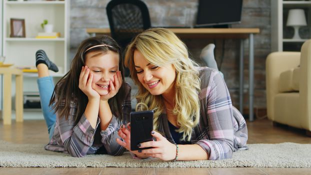 Little girl and her mother lying on the floor using smartphone for shopping.