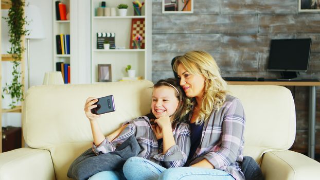 Little girl and her mother sitting on the couch in living room during a video call on the phone.