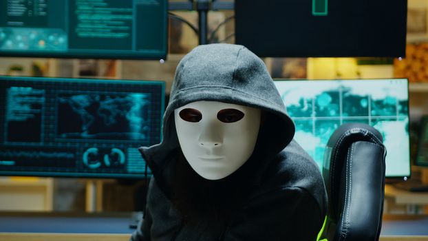 Masked hacker in his apartment looking into the camera while stealing online information.