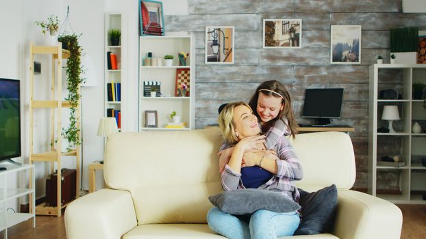 Happy little girl hugging her mother from behind while she is sitting on the couch in living room.