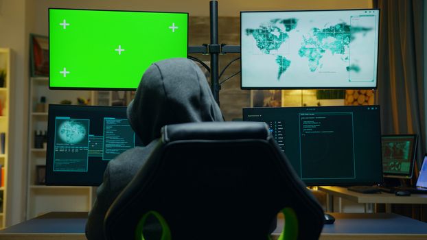 Back view of hacker wearing a hoodie using computer with green screen.