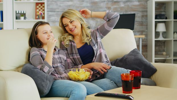 Little girl eating chips while watching movie on tv with her mother. Mother and daughter relaxing.