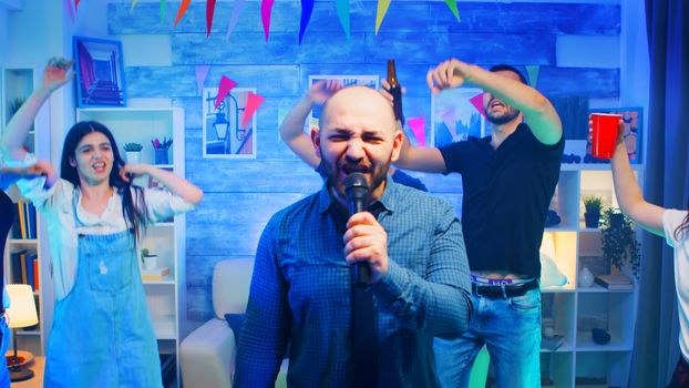 Attractive bald man doing karaoke at wild party with neon lights. Cheerful group of people.