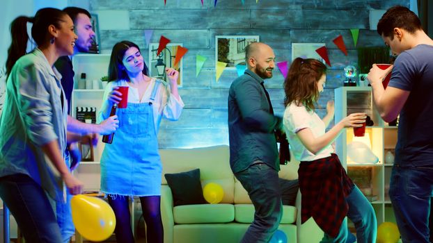 Bald attractive young man smiling while dancing at the party with his friends. Wild college party with neon lights and disco ball