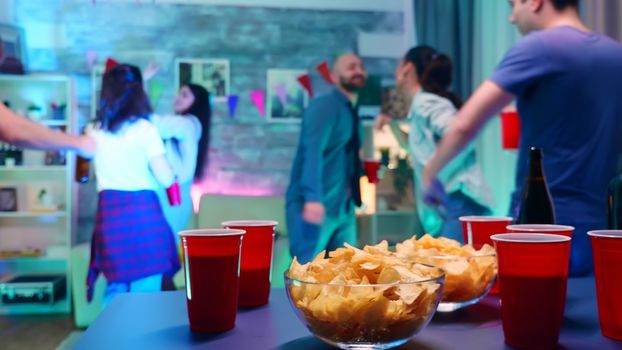 Young woman at a party with neon light taking chips from the table and a cup of beer. Young people dancing in the background at wild college party