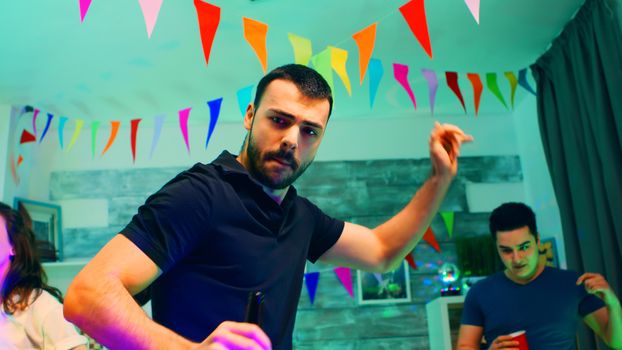 Care free young man holding a beer bottle while partying with his friends in an apartment with neon lights and disco ball