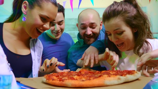 Group of young people full of excitement at arriving of pizza. Friends hangout at wild college party with neon lights