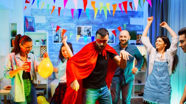 Cheerful young man wearing superhero red cap while dancing with his friends at the party in living room with neon light.