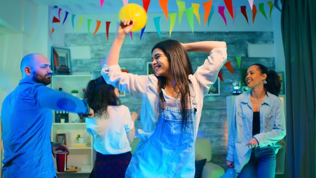 Attractive young woman partying with her friends while holding a balloon. Wild college party with a room full of neon lights, disco ball and alcohol