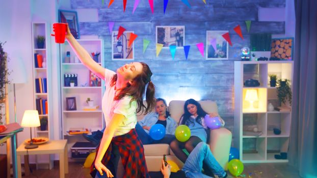 Care free young woman dancing with a cup of beer while her best friends are lying on the floor because of drinking too much alcohol.