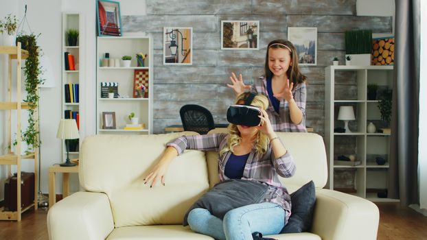 Little girl jumping around her mother while she's using virtual reality headset sitting on the couch.