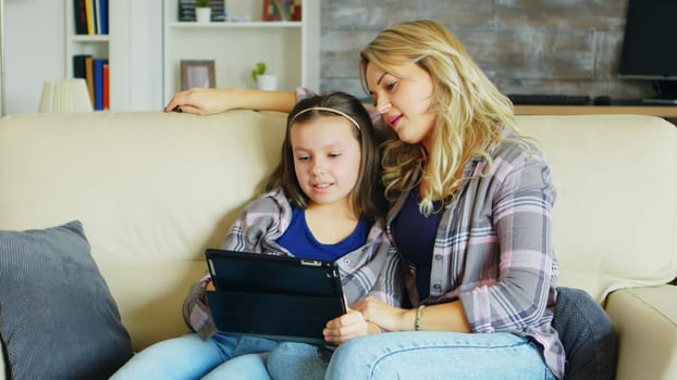 Beautiful young mother using tablet for her daughter education sitting on the couch in living room.