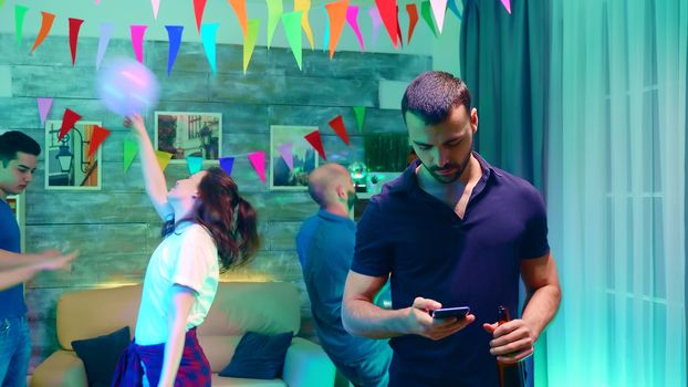 Bearded young woman holding a beer bottle while browsing bored on smartphone at the party.