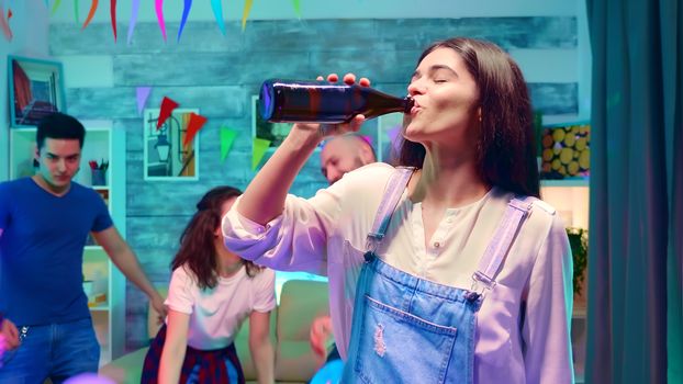 Zoom in shot of beautiful girl drinking beer from the bottle at wild party with neon lights. Group of people dancing in the background