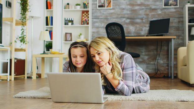 Cheerful little girl and her mother lying on the floor using laptop for shopping.