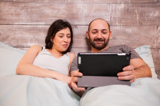 Beautiful young husband and wife wearing pajamas laughing while using tablet computer before bedtime.