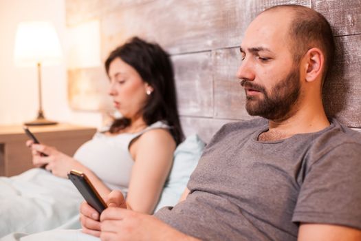 Caucasian man surfing on internet using his phone before bedtime and having a communication with his wife.