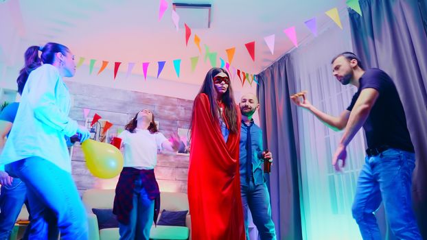 Attractive girl full of excitement wearing a superhero red cap while dancing with her friends at wild college party with neon lights.