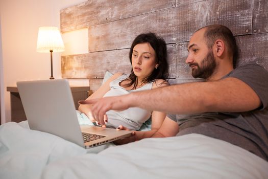 Husband pointing at laptop before bedtime. Happy married couple in pajamas.