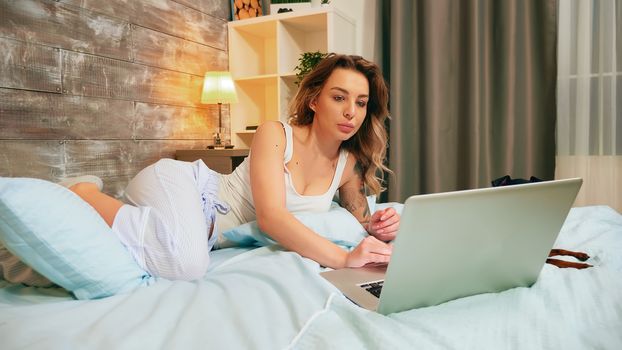 Caucasian attractive woman in pajamas lying in bed using her laptop. Happy dog.
