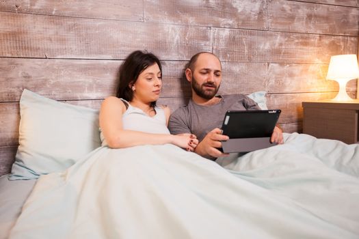 Caucasian couple wearing pajamas layibng in bed using tablet computer.