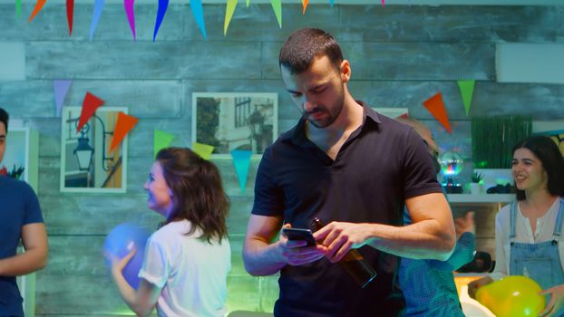 Bored millennial man using his smartphone while his friends are partying wild in the background. Room with neon lights.