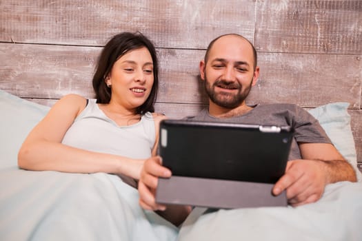 Beautiful young couple wearing pajamas sitting side by side in bed at night using tablet computer.