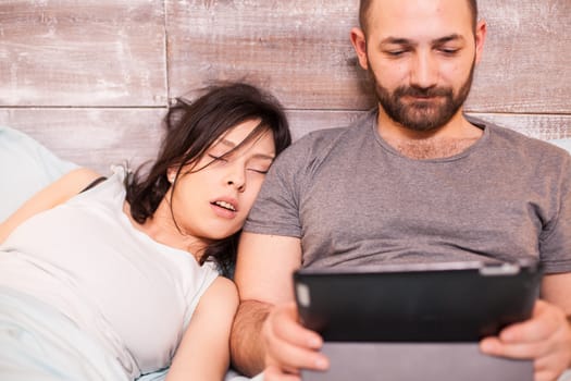 Beautiful young woman sleeping next to her husband. Man usint tablet computer.