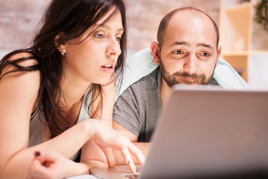 Young married couple laying in bed before sleep searching for vacation destination on laptop.