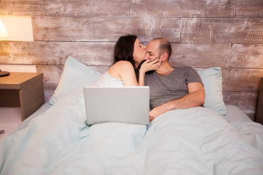 Beautiful wife in pajamas kissing her beautiful husband before bedtime. Laptop on bed.