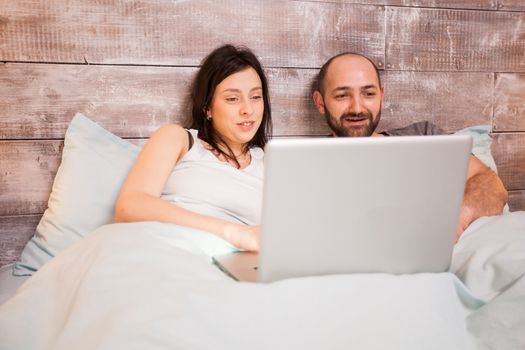 Married young couple in pajamas using laptop laying in bed before going to sleep .