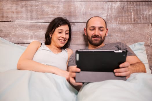 Husband and wife wearing pajamas laughing while watching a funny video on tablet computer.